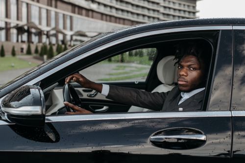 Free African American man confidently driving a sleek black luxury car outside a modern building. Stock Photo