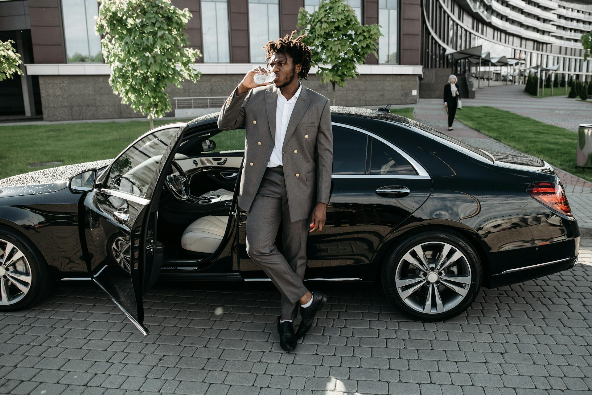 A fashionable businessman stands by a luxury car, enjoying a drink on a city street.