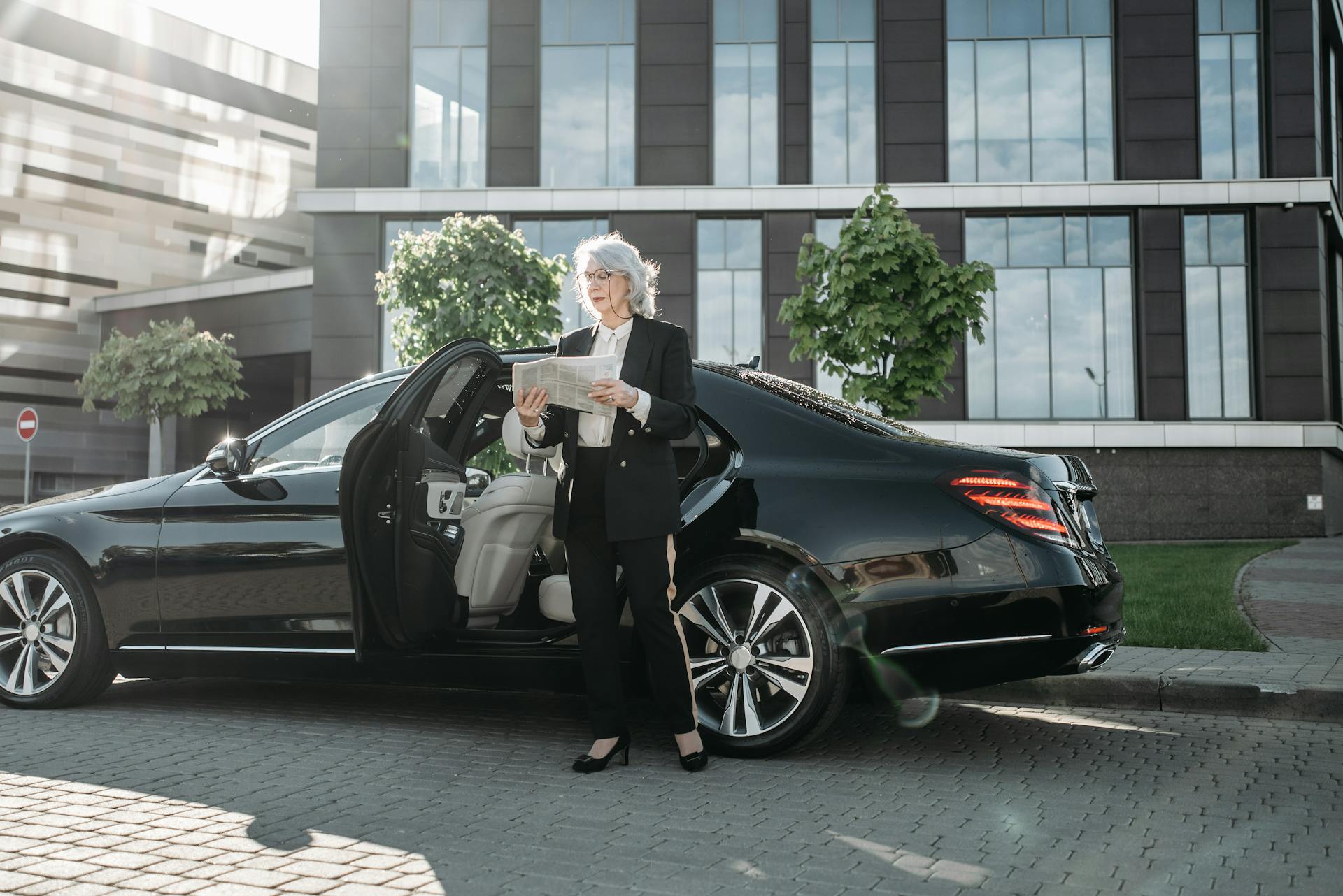 Woman in Corporate Attire Standing Beside a Luxury Car Reading Newspaper