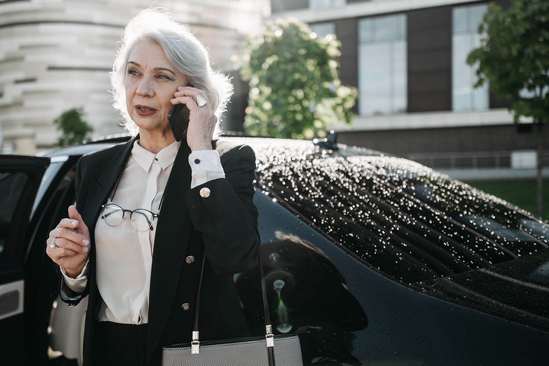 Senior woman in formal attire using a mobile phone near a black car outdoors.