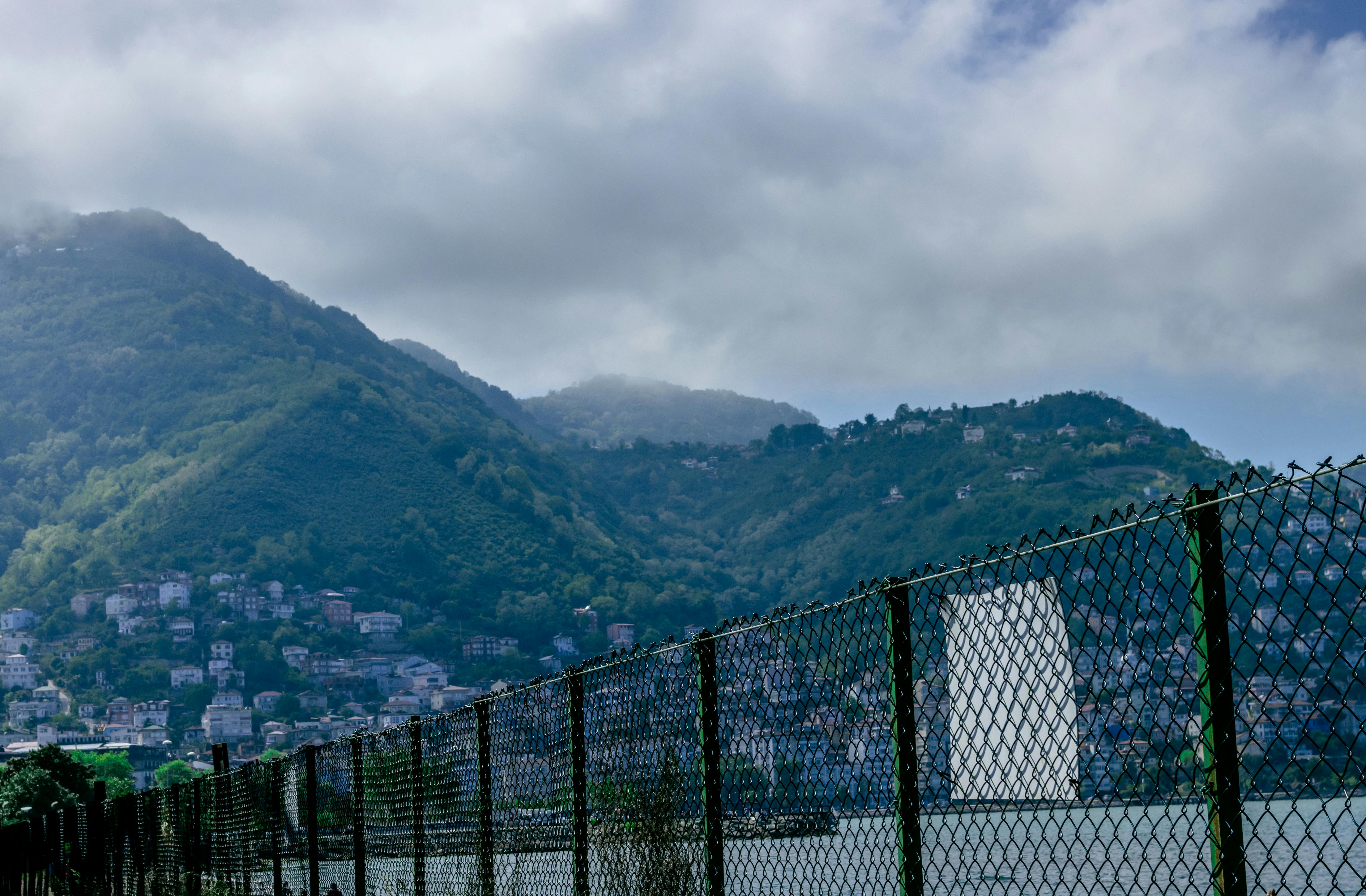 Chain Link Fence Near Body of Water and Houses on Mountainside Under Cloudy  Sky · Free Stock Photo
