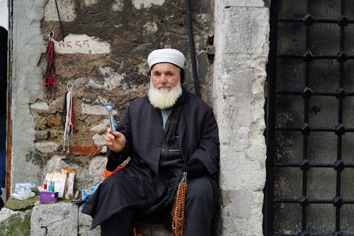 Elderly Man Wearing White Kufi Hat Sitting on Concrete Ledge