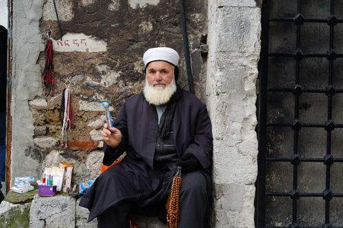 Free An elderly man in traditional clothing seated on a stone ledge, selling items in an outdoor market. Stock Photo