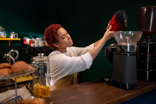 Woman barista filling coffee grinder with beans in a cozy café setting.