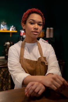 A young female barista stands confidently in a cozy café setting wearing an apron.