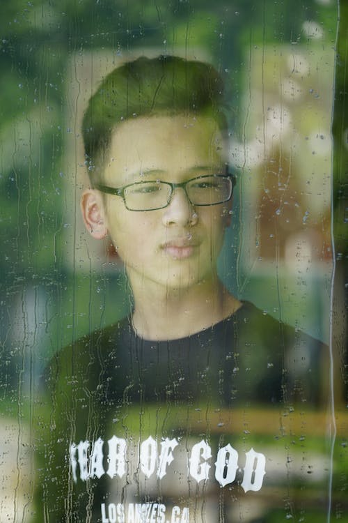 View through wet window with crystal raindrops on thoughtful ethnic teenage man in eyeglasses looking away