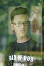 View through wet window with crystal raindrops on thoughtful ethnic teenage man in eyeglasses looking away