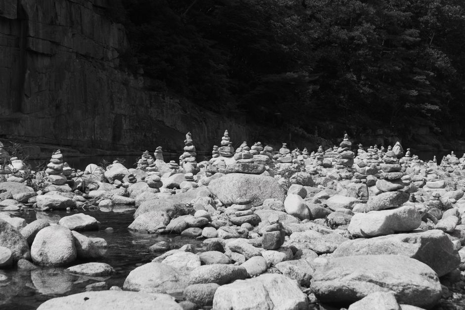 Black and white image of stacked stone cairns by a river in nature's calm setting.