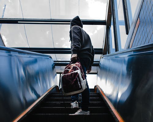 Persona Vestida Con Chaqueta Con Capucha Negra De Pie En La Escalera Mecánica Mientras Sostiene La Mochila