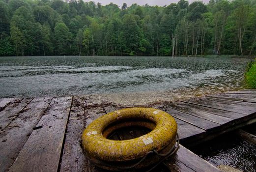 A serene view of a lakeside wooden dock with a lifebuoy under gentle rainfall.
