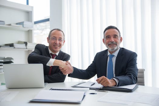 Two businessmen in formal suits shaking hands across a table in an office setting.