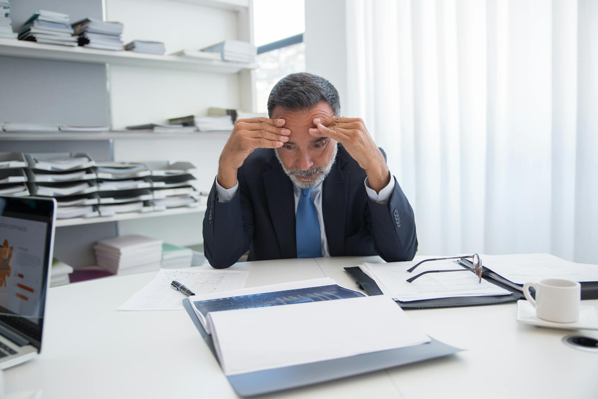 An elderly businessman in a suit appears stressed while sitting at a desk with documents in an office.