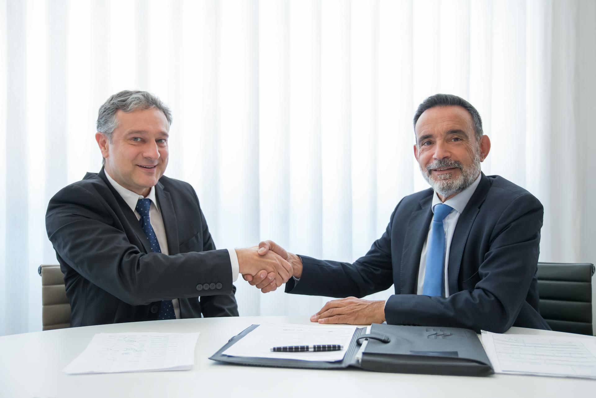 Two businessmen in suits shaking hands at an office table with documents.