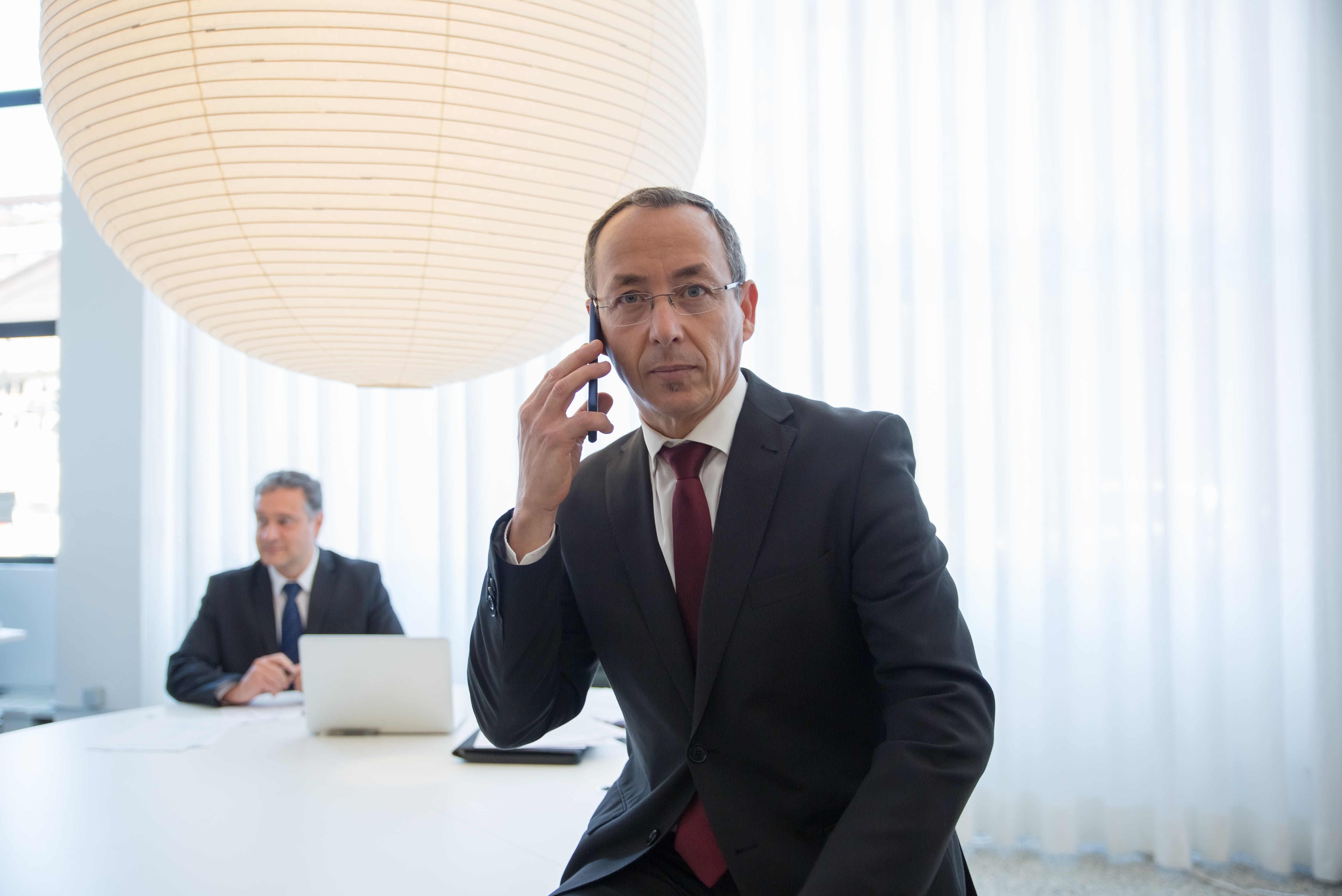 A businessman in a suit making a phone call during a formal meeting in a modern, bright office.