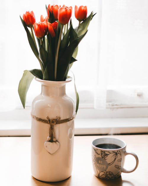一杯のコーヒーの横にある白いセラミック花瓶の赤いチューリップの花