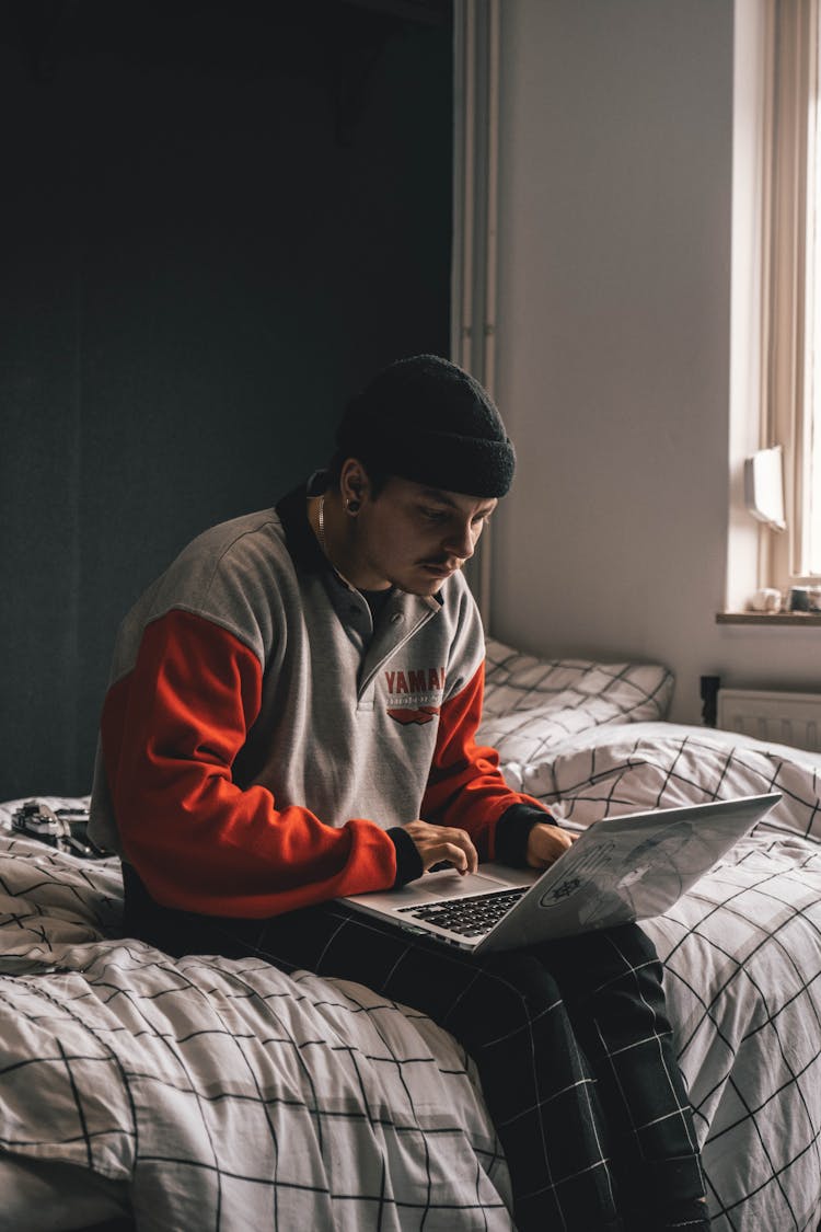 A Man In Gray And Red Sweater Using His Laptop While Sitting On The Bed