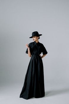 Stylish adult woman in a long black dress and hat posing confidently indoors.