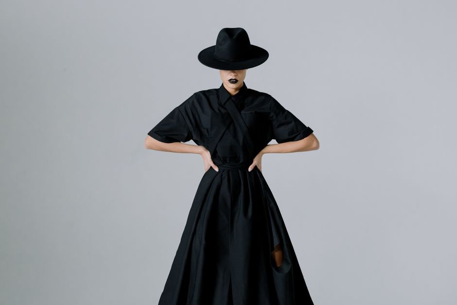Elegant model in a black dress with a hat, exuding a mysterious and fashionable vibe in a studio portrait.