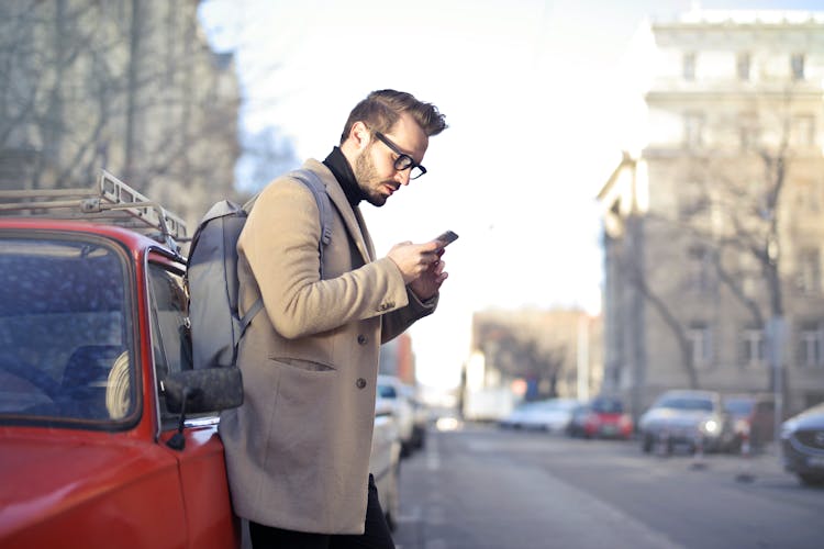 Man In Beige Coat Holding Phone Leaning On Red Vehicle