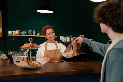 Free A friendly barista hands over pastries to a customer at a cozy café counter. Warm ambiance indoors. Stock Photo