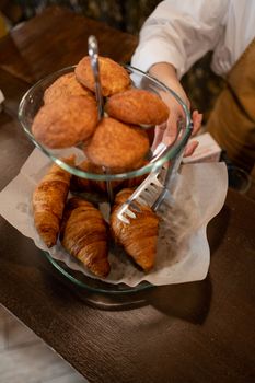 Croissants and muffins on a glass stand held with tongs, presented on a wooden table.