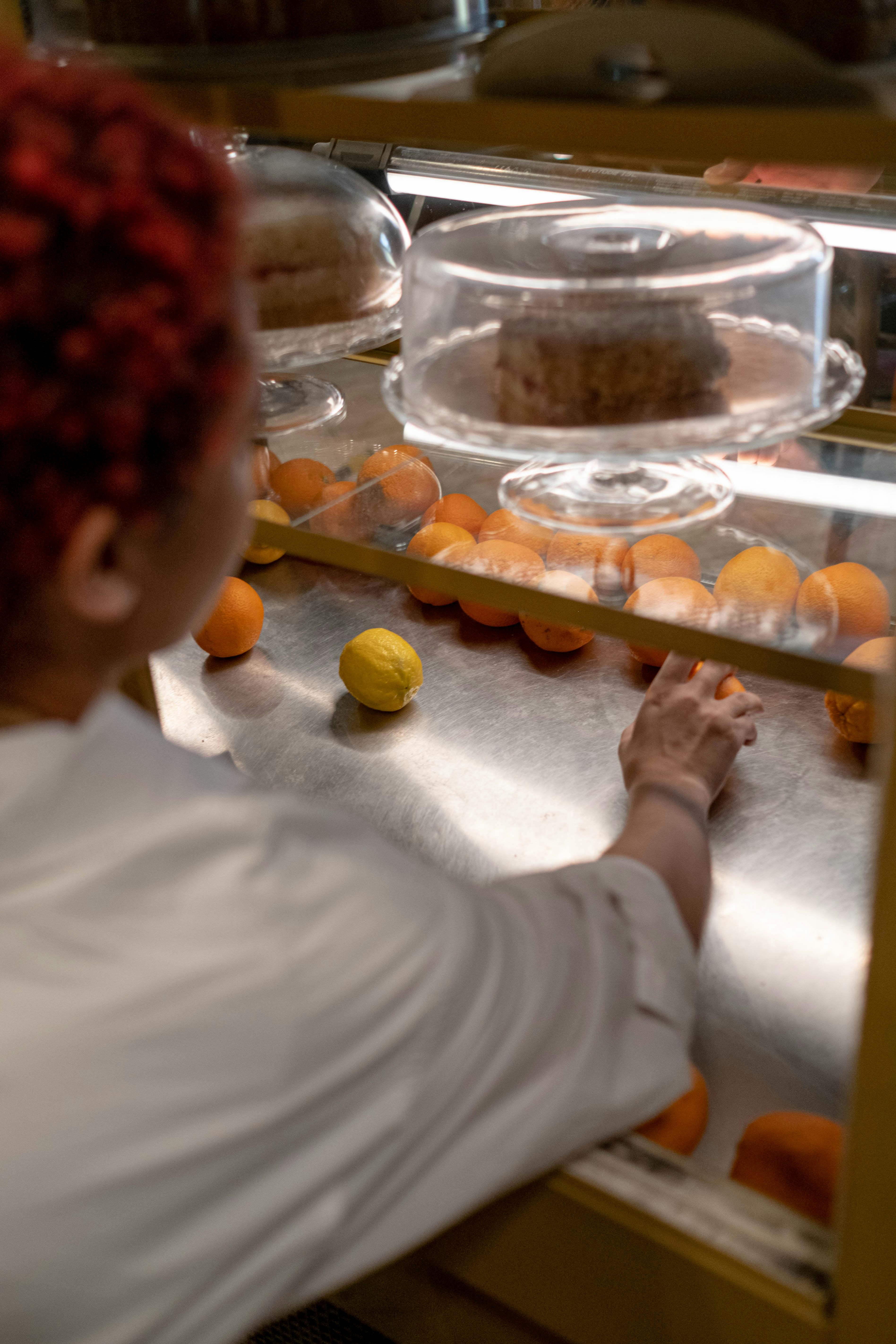 A person reaching for oranges in a café display, with cakes overhead.