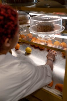 A person reaching for oranges in a café display, with cakes overhead.