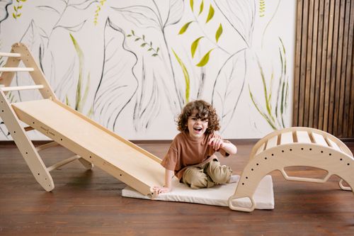 Free A happy child with curly hair enjoys playing on a wooden slide in a modern playroom. Stock Photo