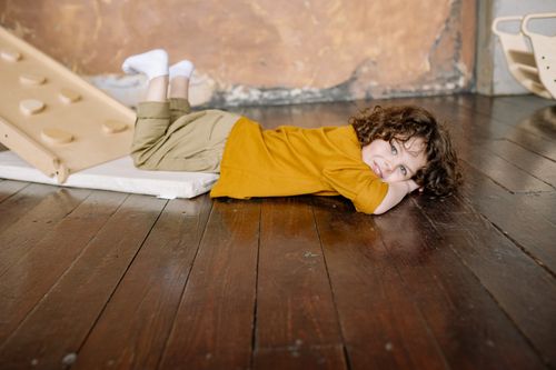 Free Smiling curly-haired child enjoying playtime indoors, lying on a wooden floor. Stock Photo