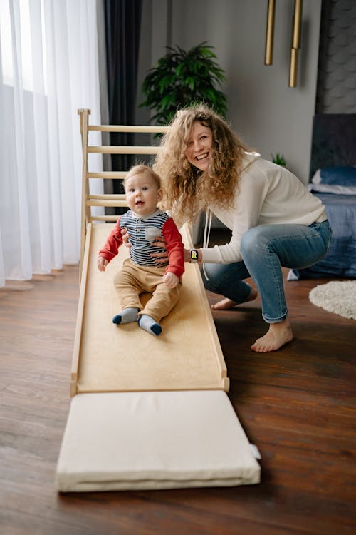 A Woman in White Long Sleeves Holding Her Baby Boy Sitting on a Wooden Slide