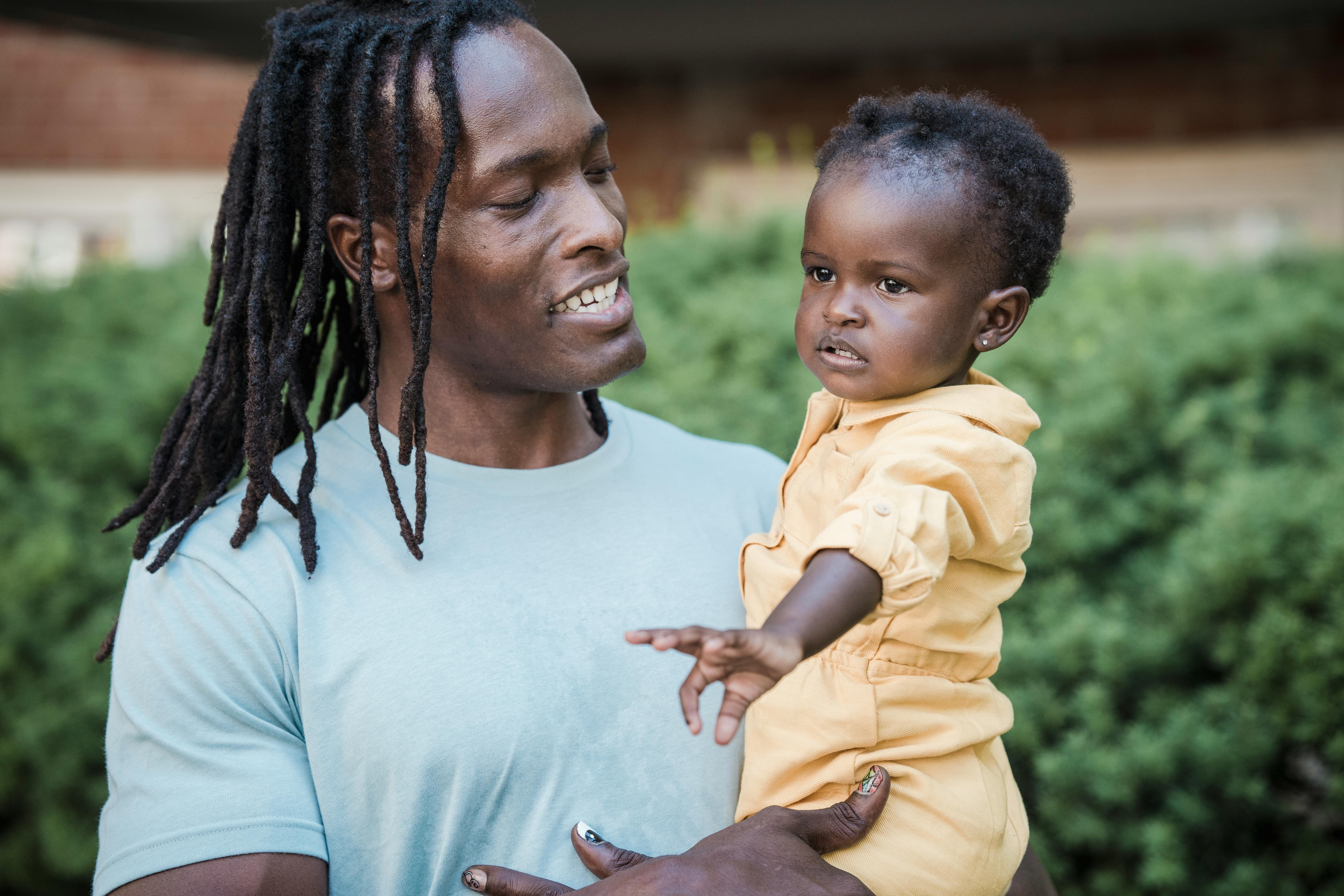 A father lovingly holding his daughter in a sunlit park, both smiling warmly.