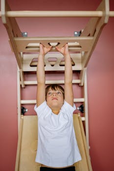 Young boy in white t-shirt hanging on indoor climbing equipment, pink wall backdrop.