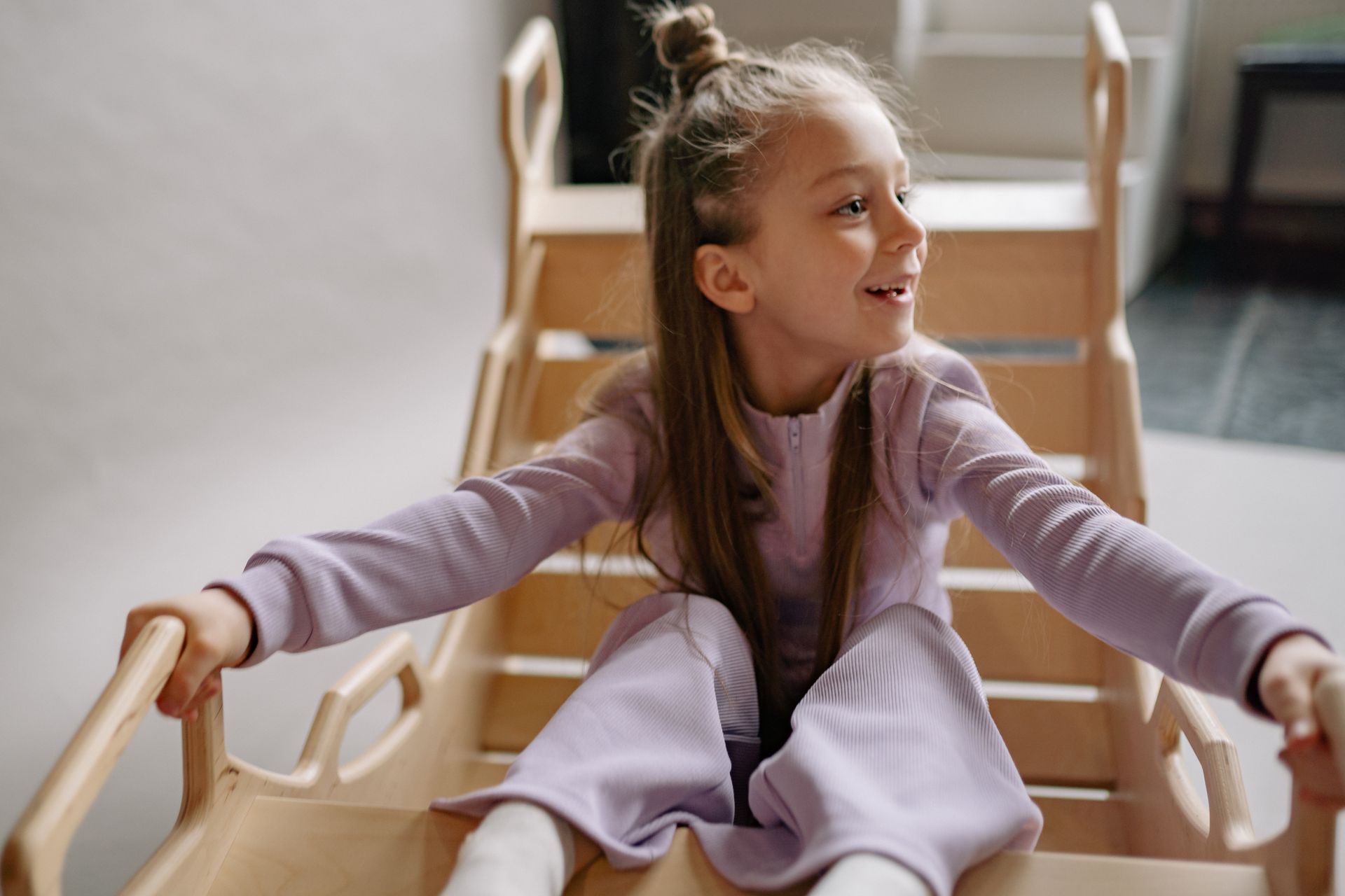 A young girl in lilac clothing happily playing on a wooden slide indoors, smiling brightly.