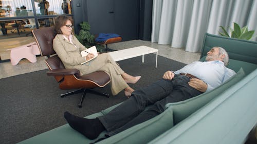 Professional Woman Writing Notes in Front of a Man Lying on Sofa