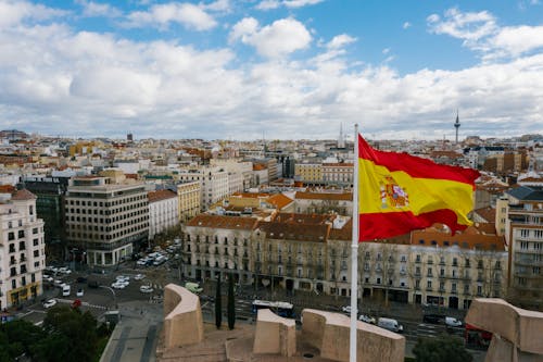 Drone view of Spanish city with aged buildings and national flag under cloudy blue sky