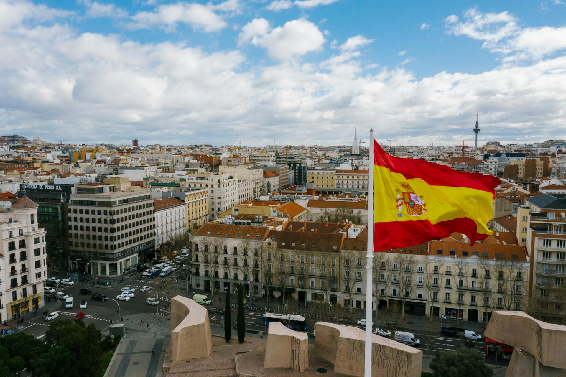 Drone view of Spanish city with aged buildings and national flag under cloudy blue sky