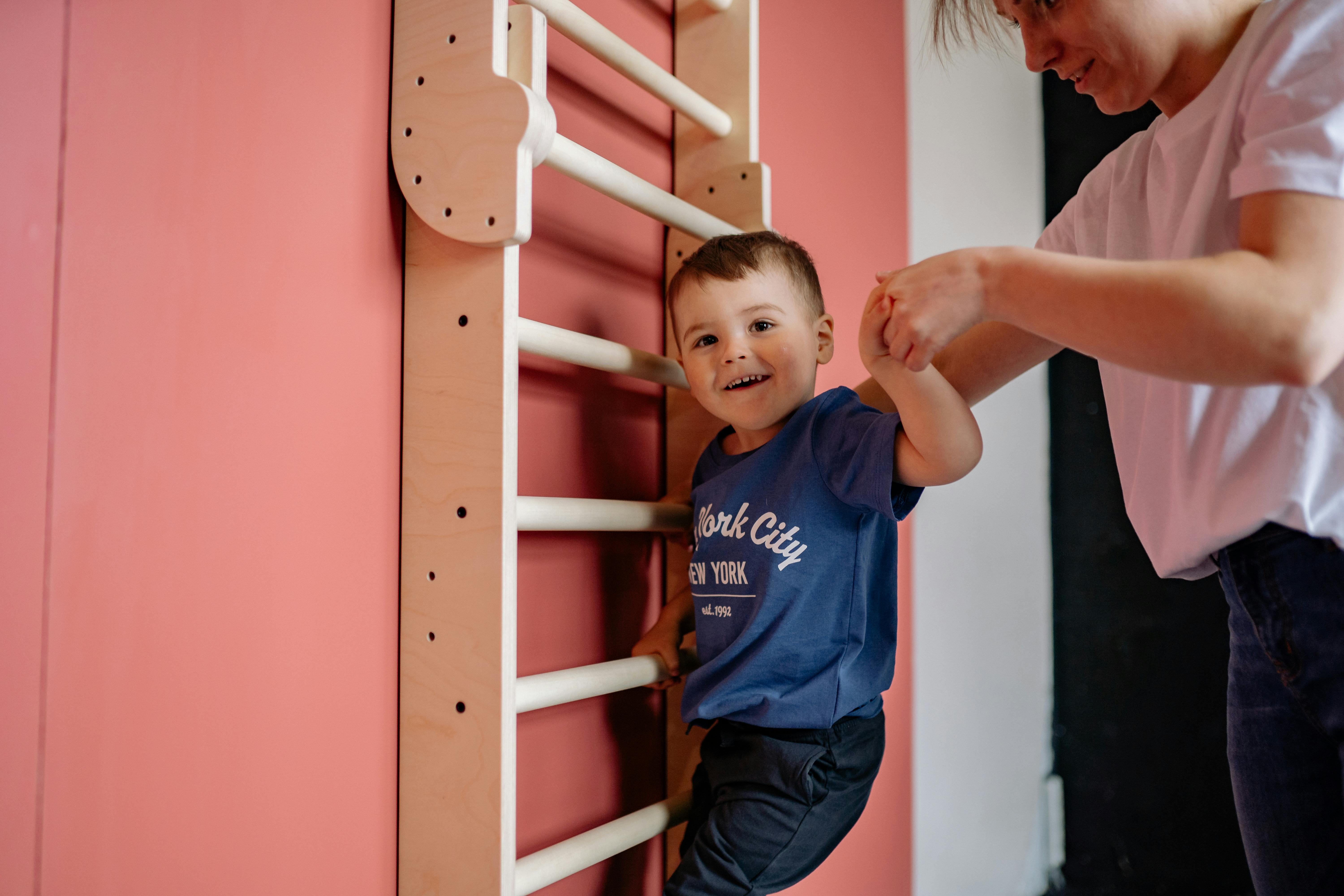 A smiling boy climbs a wooden ladder with a woman's assistance, embodying teamwork and joy indoors.