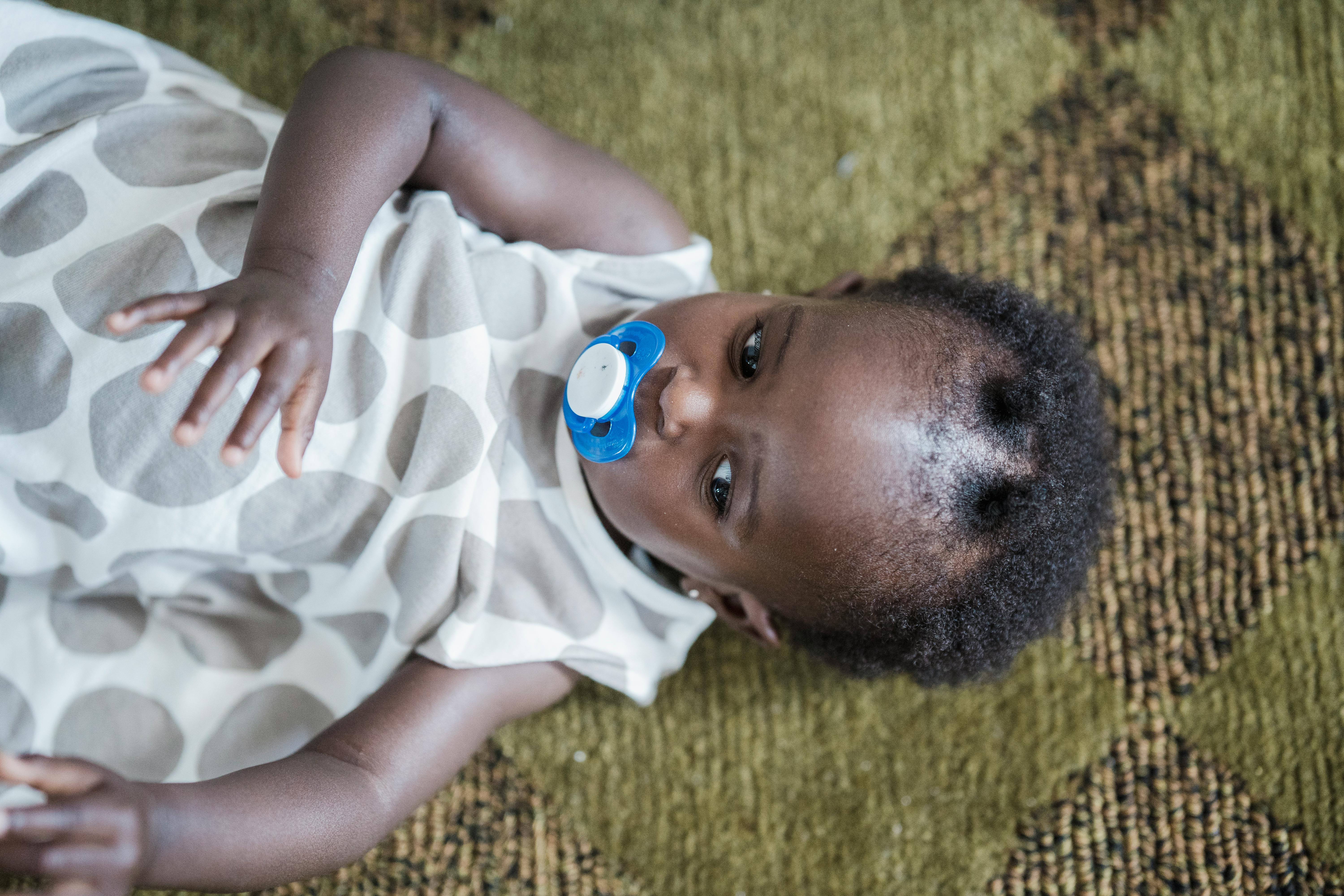 Cute baby girl lying on carpet, afro hair with pacifier, showcasing innocence and charm.