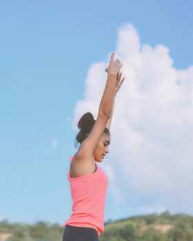 A young woman in a tank top performing a yoga pose outdoors on a sunny day, raising her arms against a clear sky.