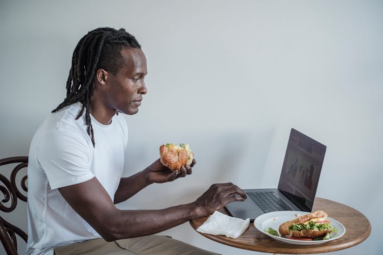 A Man Using A Laptop While Eating A Sandwich