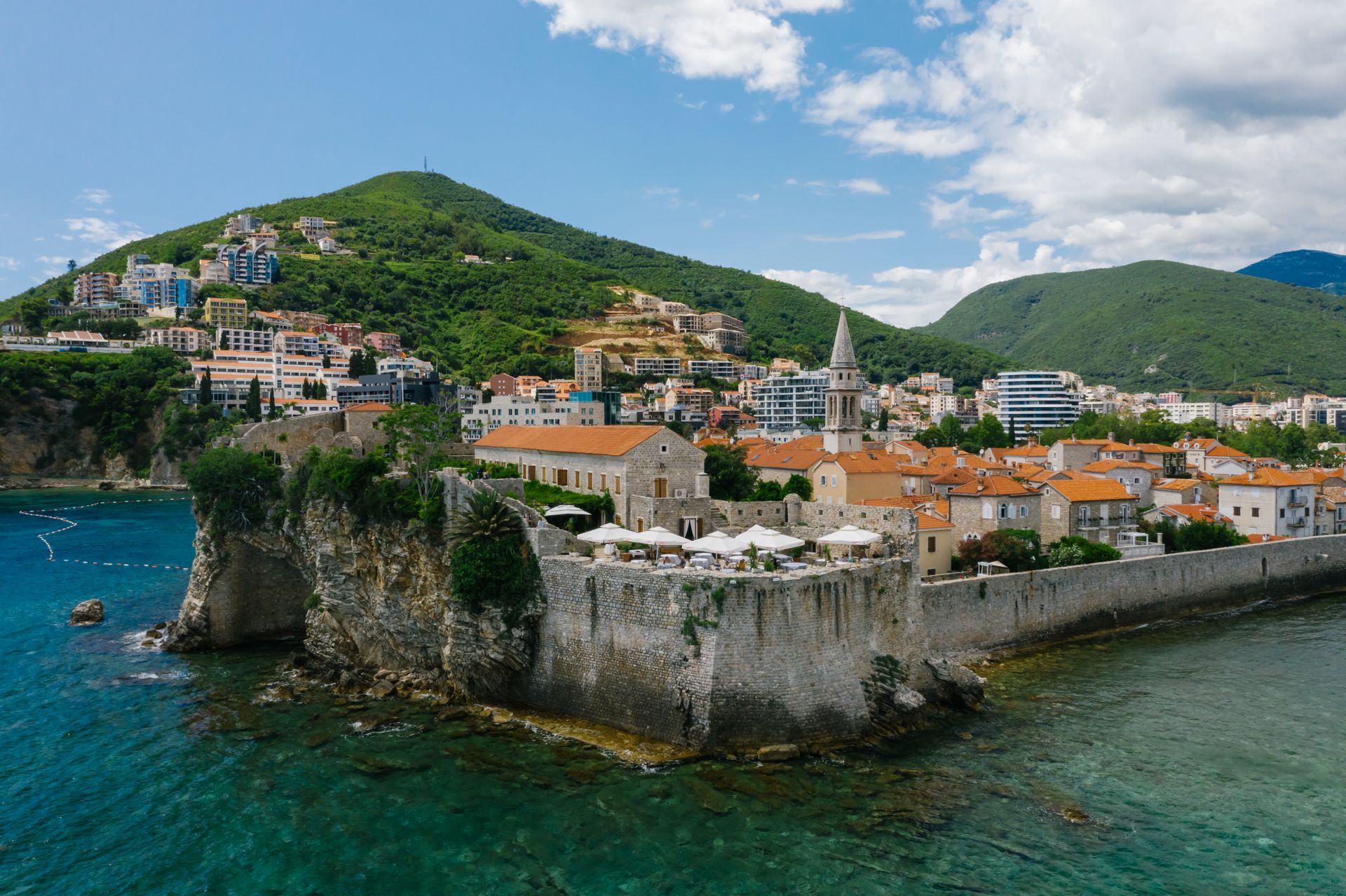 Aerial view of a historic town on the Adriatic coast, featuring old stone walls and lush green mountains.