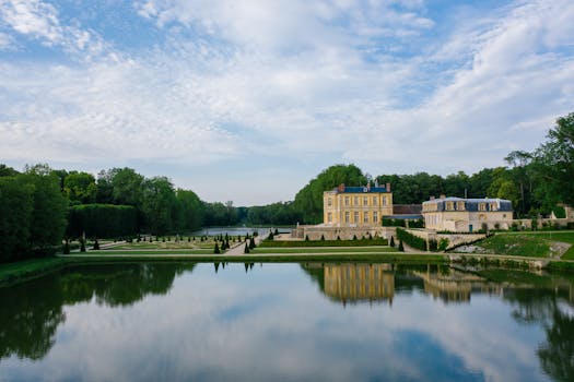 Scenic view of a French chateau surrounded by greenery and a reflective lake under a blue sky.