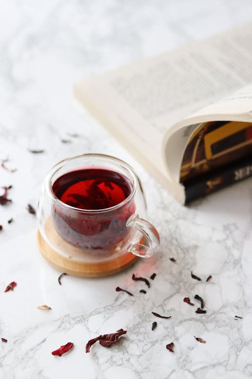 High angle of cup on wooden board full of freshly brewed hot tea placed near opened book with soft cover lying on table with light surface