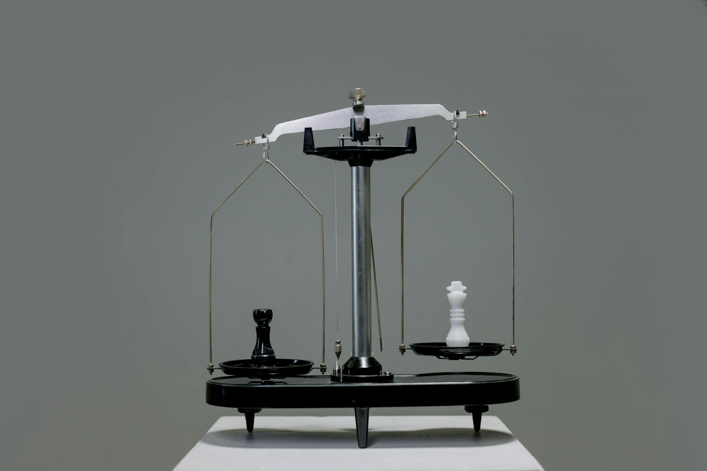 Balance scales with a black "queen" chess piece on the left and a white "king" chess piece on the right. The white "king" chess piece is higher up
