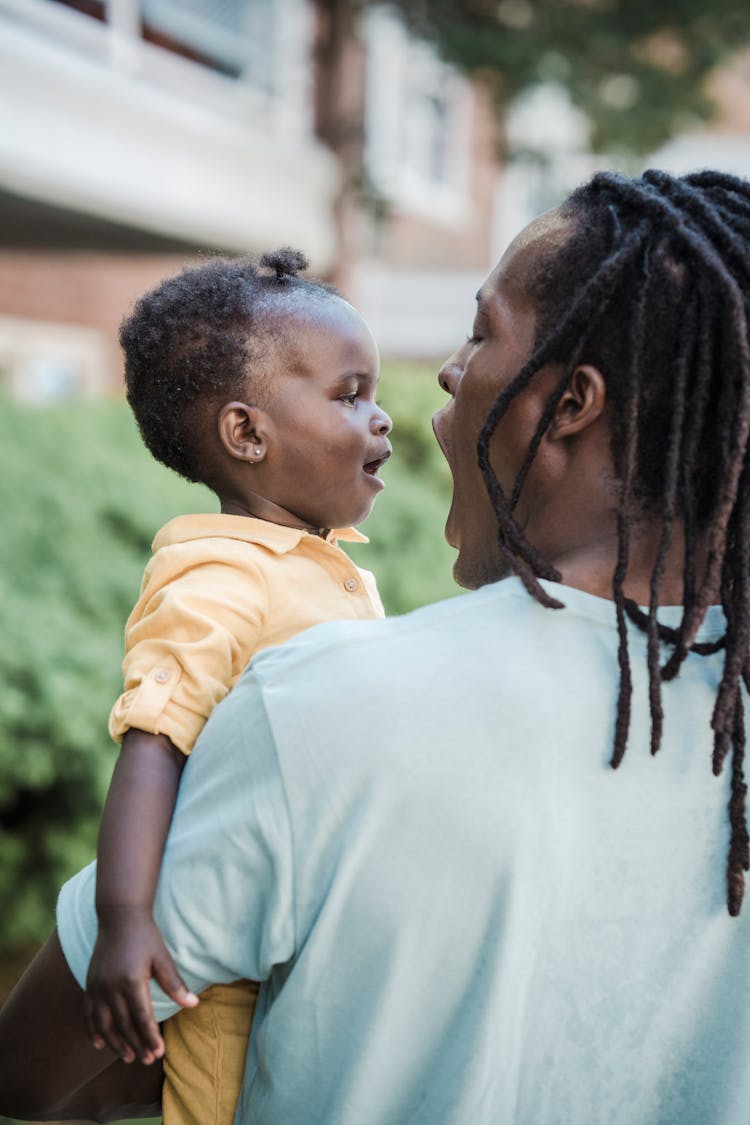 Man With Dreadlocks Holding A Baby Girl In Arms And Making Faces In A Residential Area