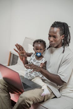 A loving father spends quality time with his child on a sofa, browsing on a laptop.