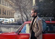 阳光明媚的布达佩斯街道上，一位戴眼镜、背背包的时尚男子靠在一辆老式红色汽车上