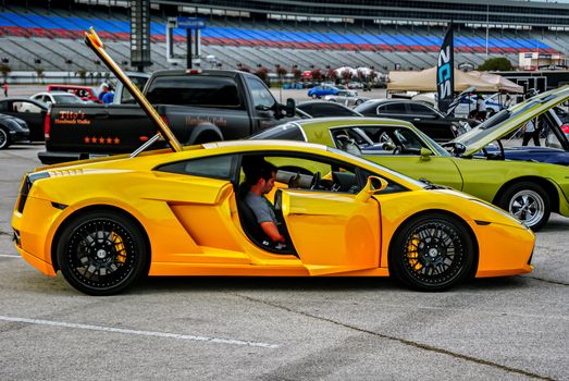 Yellow sports car displayed at an outdoor car show with open doors and other vehicles in the background.
