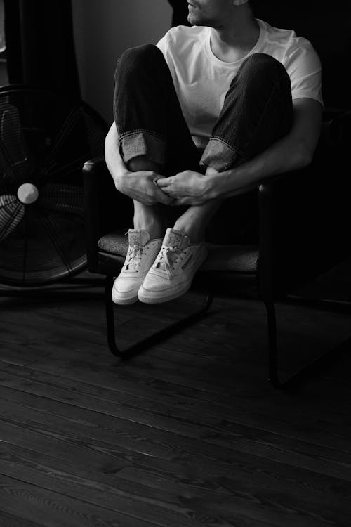 Black and white of crop anonymous male in sneakers sitting on chair placed on wooden floor and embracing knees