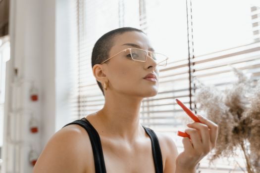 A stylish woman with eyeglasses applying lipstick confidently indoors, showcasing her unique fashion sense.